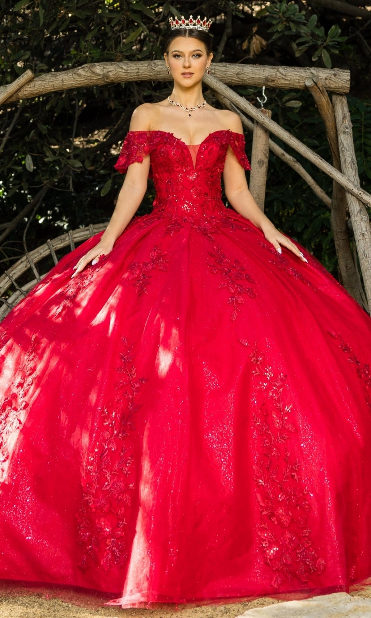 A woman in the Quinceanera Ball Gown 8045J, a red off-the-shoulder dress with glitter lace embroidery and a crown, stands outdoors before wooden branches.